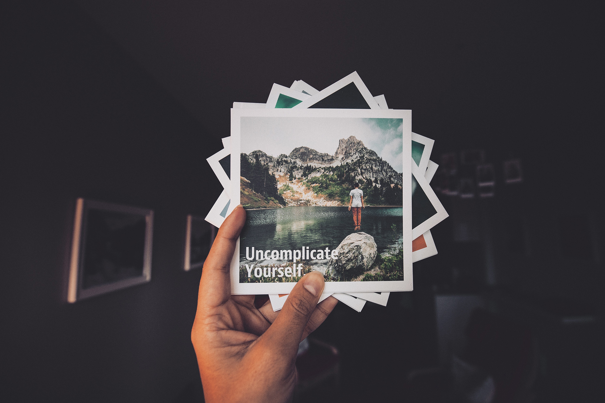 image of hand holding stack of photographs. Top photo is of a woman standing in front of a serene landscape, with the words, "Uncomplicate Yourself"